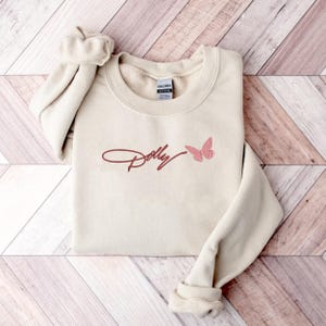 May include: A sand-colored crewneck sweatshirt featuring the word Dolly embroidered in a dark red cursive script, accompanied by a small, light pink embroidered butterfly design positioned to the right of the text on the chest area.