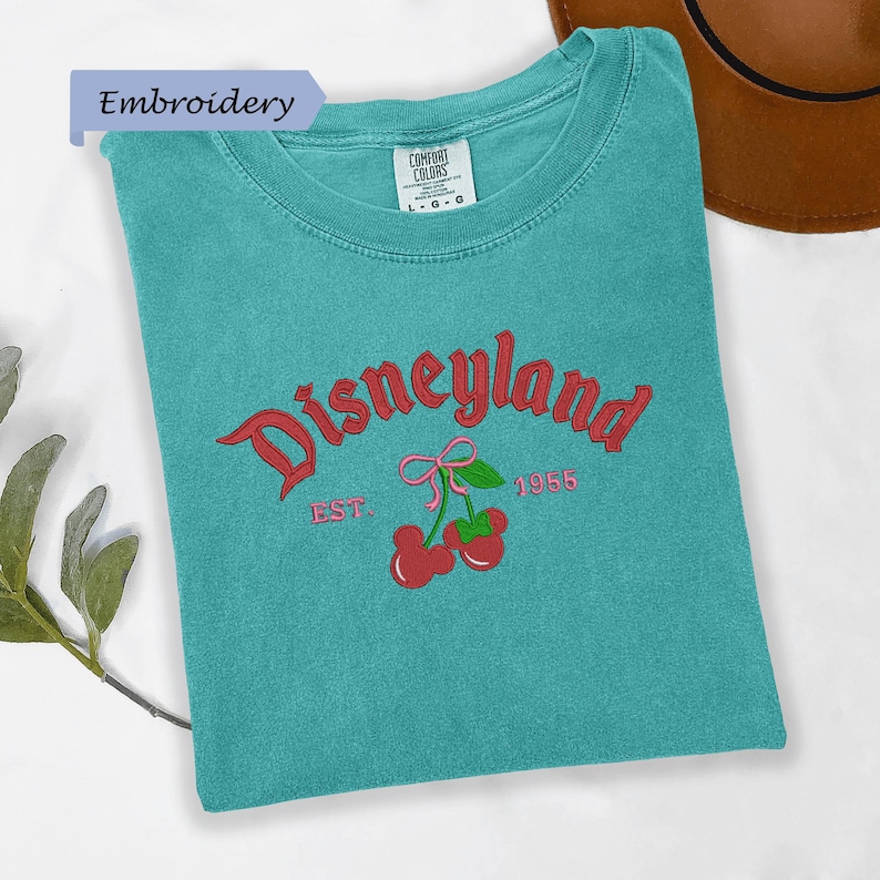 A turquoise t-shirt with a Disneyland logo and text, along with a brown hat, is displayed on a white surface.