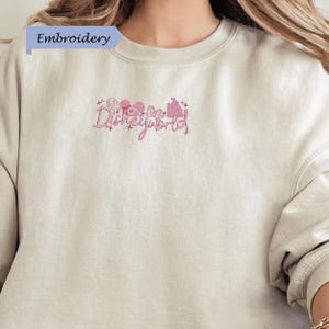 A close-up of a person wearing a beige sweater with a pink embroidered logo that says "Disneyworld".