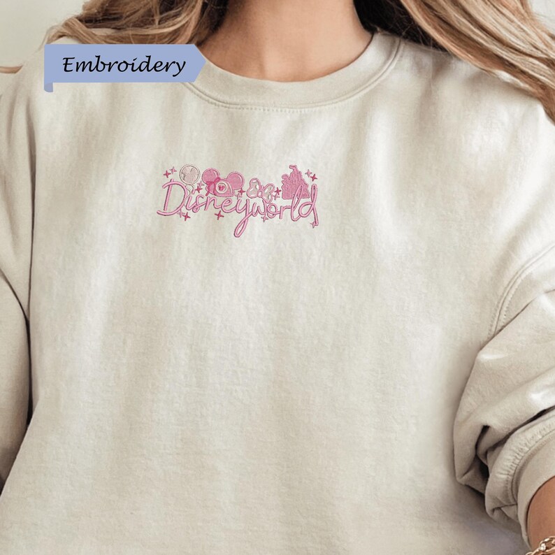 A close-up of a person wearing a beige sweater with a pink embroidered logo that says "Disneyworld".