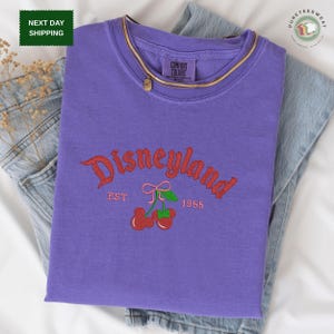A purple t-shirt with the text "Disneyland EST. 1955" printed on it, along with a graphic of cherries. The t-shirt is placed on a white surface, and there is a pair of jeans visible in the background.