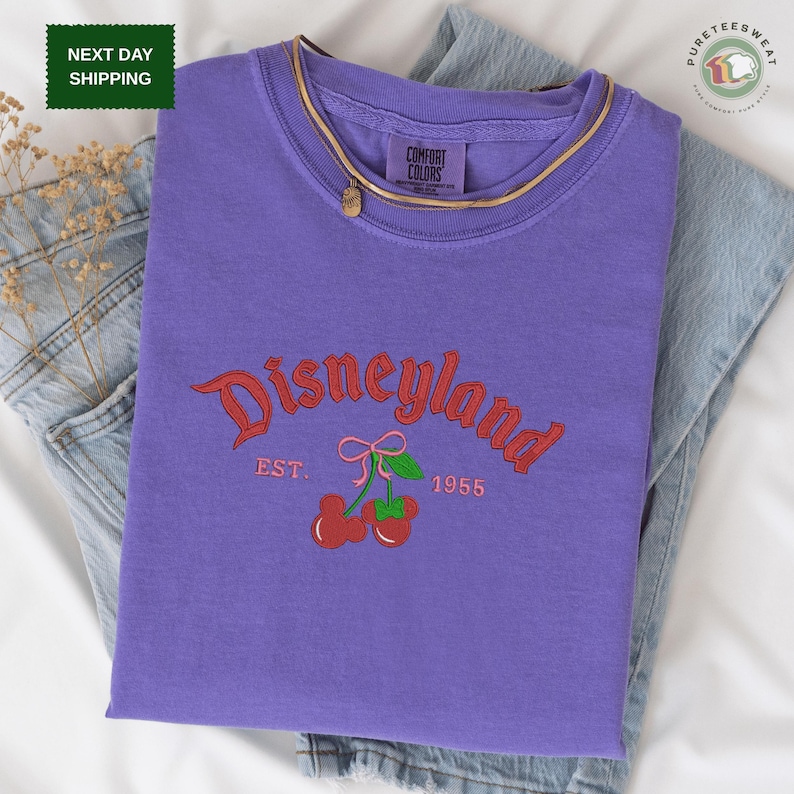 A purple t-shirt with the text "Disneyland EST. 1955" printed on it, along with a graphic of cherries. The t-shirt is placed on a white surface, and there is a pair of jeans visible in the background.