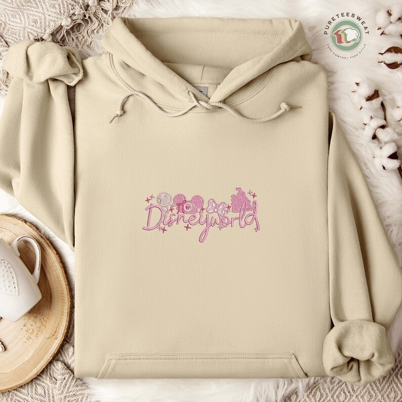 A beige hoodie with the text "Disneyworld" embroidered on it, along with various Disney-themed illustrations, is displayed on a bed with a white blanket and a white teapot.