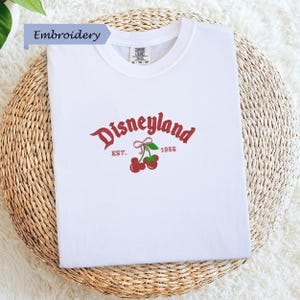 A white t-shirt with a red and green embroidered logo that says "Disneyland EST. 1955" and features a red ribbon and cherries.
