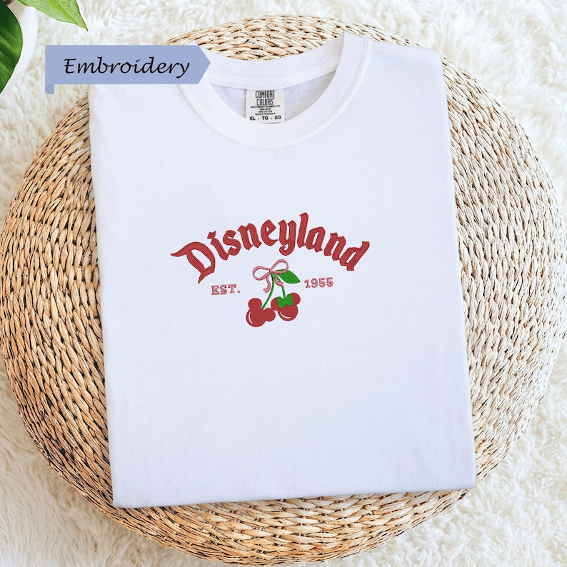 A white t-shirt with a red and green embroidered logo that says "Disneyland EST. 1955" and features a red ribbon and cherries.