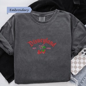 A gray t-shirt with a red and green embroidered design that says "Disneyland EST. 1955" and features a red bow and cherries.