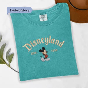 A turquoise t-shirt with the text "Disneyland EST. 1955" and a Mickey Mouse graphic, along with a brown hat, is displayed on a white surface.