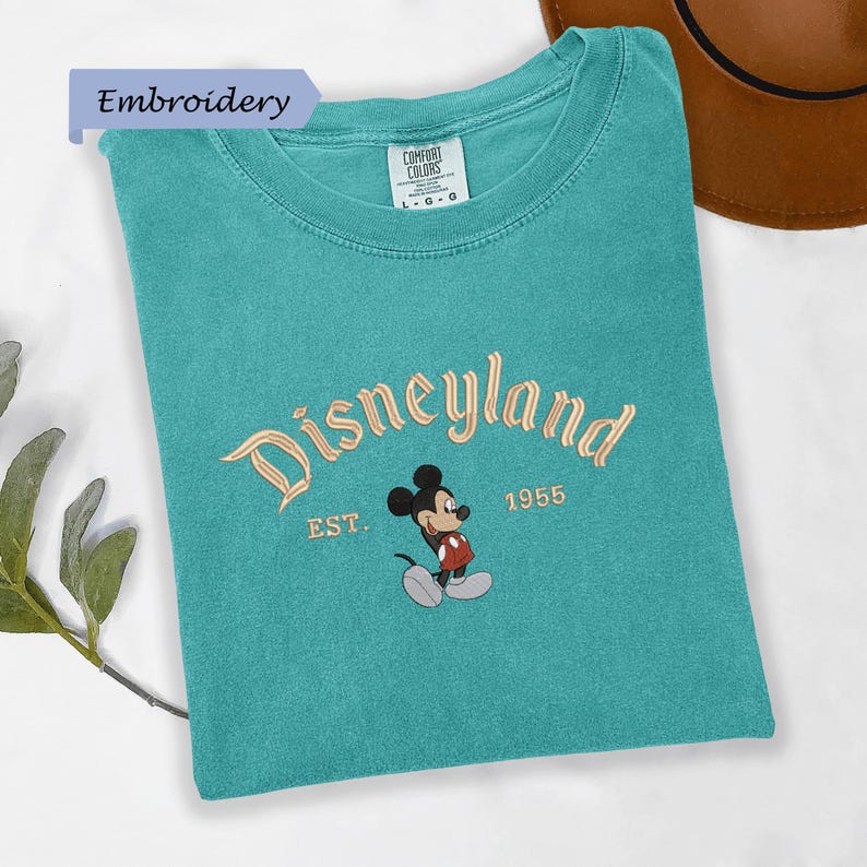 A turquoise t-shirt with the text "Disneyland EST. 1955" and a Mickey Mouse graphic, along with a brown hat, is displayed on a white surface.