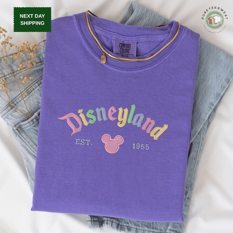 A purple t-shirt with the text "Disneyland EST. 1955" printed on it, along with a Mickey Mouse logo, is displayed on a bed.