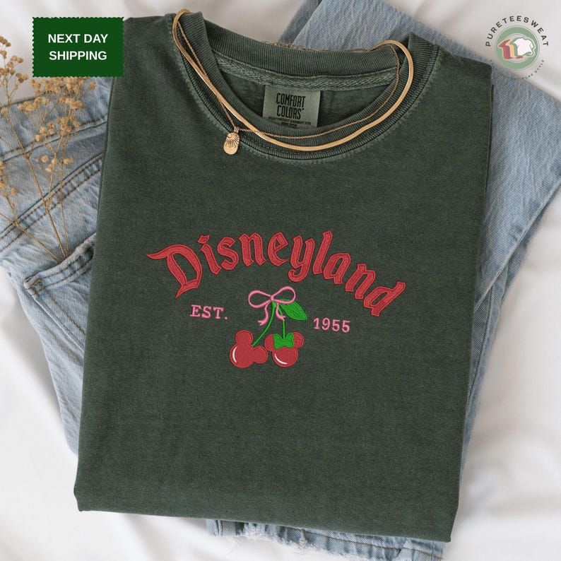A green t-shirt with a red and green design featuring the text "Disneyland EST. 1955" and cherries, laid out on a white surface.