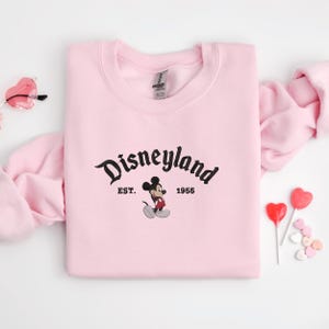 A pink sweatshirt with the text "Disneyland EST. 1955" and a Mickey Mouse graphic, along with a pair of pink glasses and heart-shaped lollipops, are arranged on a white background.