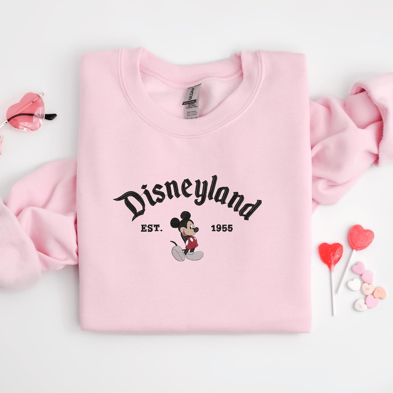 A pink sweatshirt with the text "Disneyland EST. 1955" and a Mickey Mouse graphic, along with a pair of pink glasses and heart-shaped lollipops, are arranged on a white background.