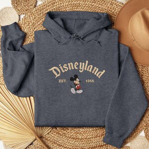 A gray hoodie with the Disneyland logo and the year 1955 printed on it, along with a straw hat and a fan, all laid out on a woven mat.