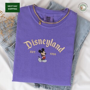 A purple t-shirt with the text "Disneyland EST. 1955" printed on it, along with a Mickey Mouse graphic. The t-shirt is laid out on a white surface, with a pair of jeans visible in the background.