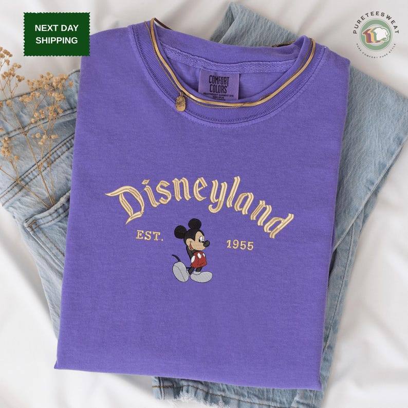 A purple t-shirt with the text "Disneyland EST. 1955" printed on it, along with a Mickey Mouse graphic. The t-shirt is laid out on a white surface, with a pair of jeans visible in the background.