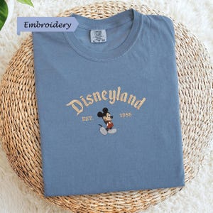 A blue t-shirt with a Mickey Mouse embroidered on it, placed on a woven basket.