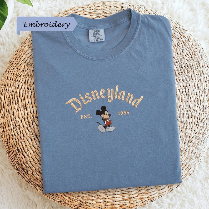 A blue t-shirt with a Mickey Mouse embroidered on it, placed on a woven basket.