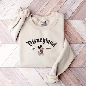 A beige sweatshirt with the text "Disneyland EST. 1955" and a Mickey Mouse graphic printed on it, laid out on a wooden surface.
