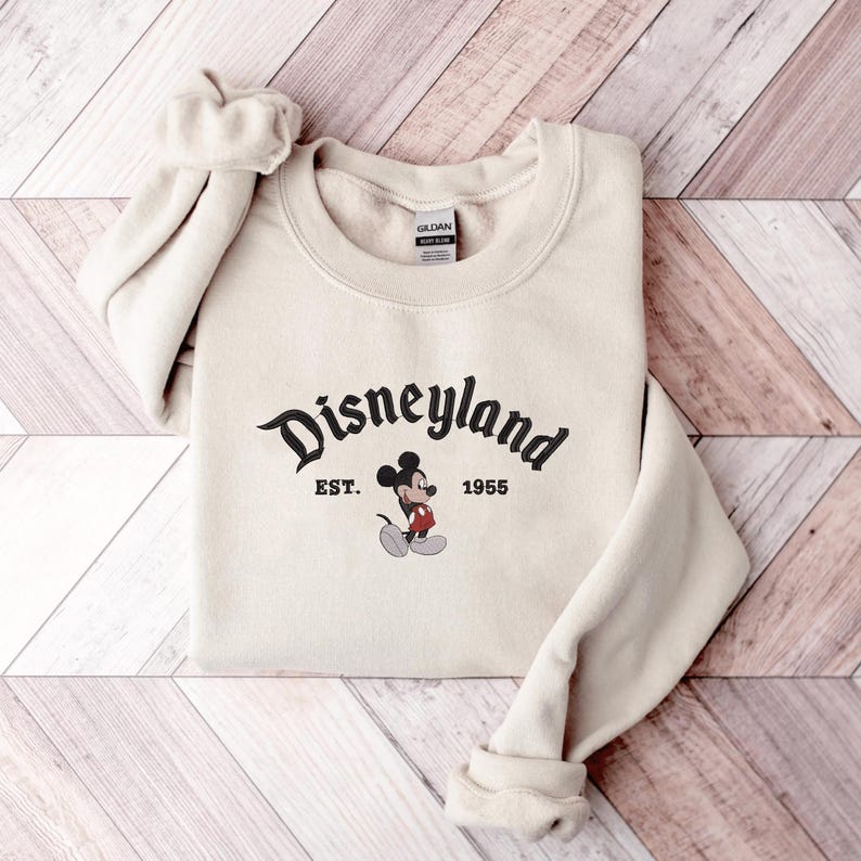 A beige sweatshirt with the text "Disneyland EST. 1955" and a Mickey Mouse graphic printed on it, laid out on a wooden surface.
