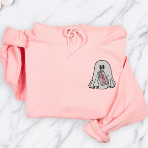 May include: A light pink hoodie with a drawstring hood and long sleeves. The hoodie features a small embroidered design of a ghost holding a Diet Coke can. The ghost is gray, and the can is red and white.