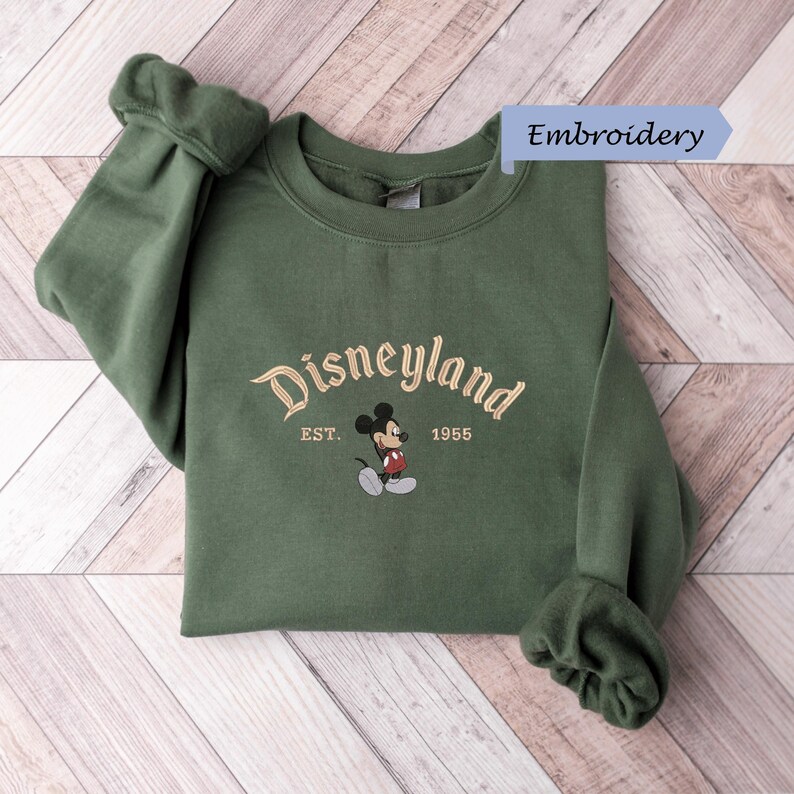 A green sweatshirt with the text "Disneyland EST. 1955" and a Mickey Mouse embroidered on it, placed on a wooden surface.