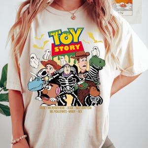 May include: Cream-coloured t-shirt featuring a colourful graphic of Toy Story characters in skeleton costumes. The design includes the Toy Story logo and character names. A fun, Halloween-themed tee.
