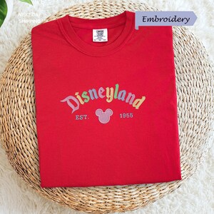 A red t-shirt with the text "Disneyland EST. 1955" embroidered on it, along with a Mickey Mouse logo, is displayed on a woven basket.
