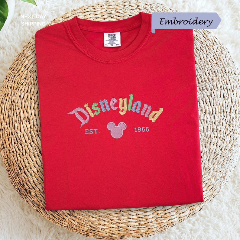 A red t-shirt with the text "Disneyland EST. 1955" embroidered on it, along with a Mickey Mouse logo, is displayed on a woven basket.