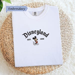 A white t-shirt with the text "Disneyland EST. 1955" and a Mickey Mouse graphic printed on it, placed on a woven basket.