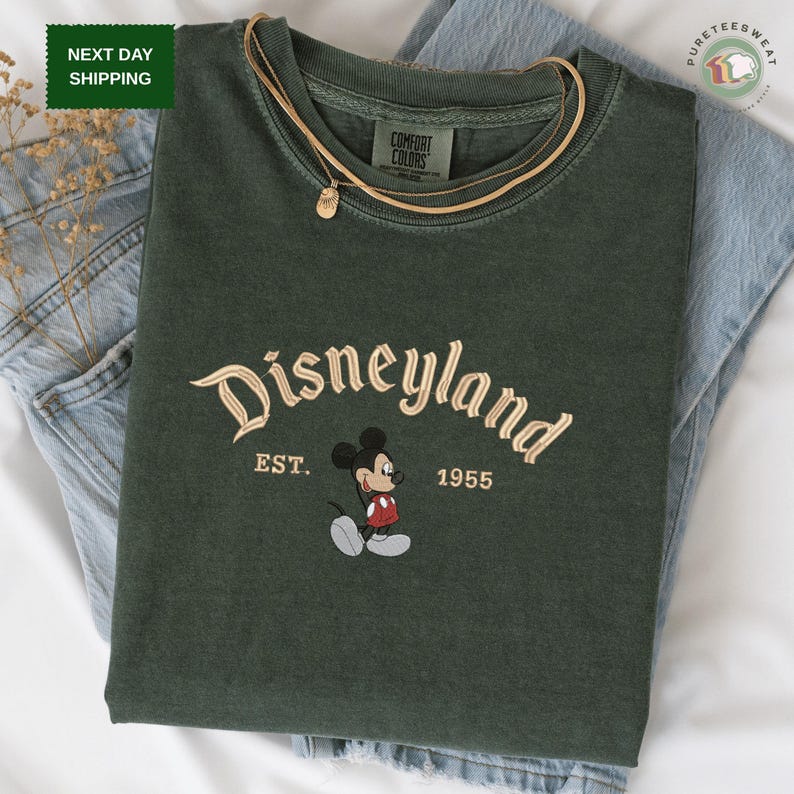 A green t-shirt with the text "Disneyland EST. 1955" printed on it, along with a Mickey Mouse graphic. The t-shirt is placed on top of a pair of blue jeans.