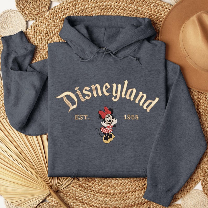 A gray hoodie with the text "Disneyland EST. 1955" and a Minnie Mouse graphic, along with a straw hat, a fan, and a woven mat, all arranged on a woven surface.