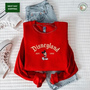 A red Disneyland sweatshirt with the text "Disneyland EST. 1955" printed on it, along with a pair of jeans and white sneakers, all laid out on a white surface.
