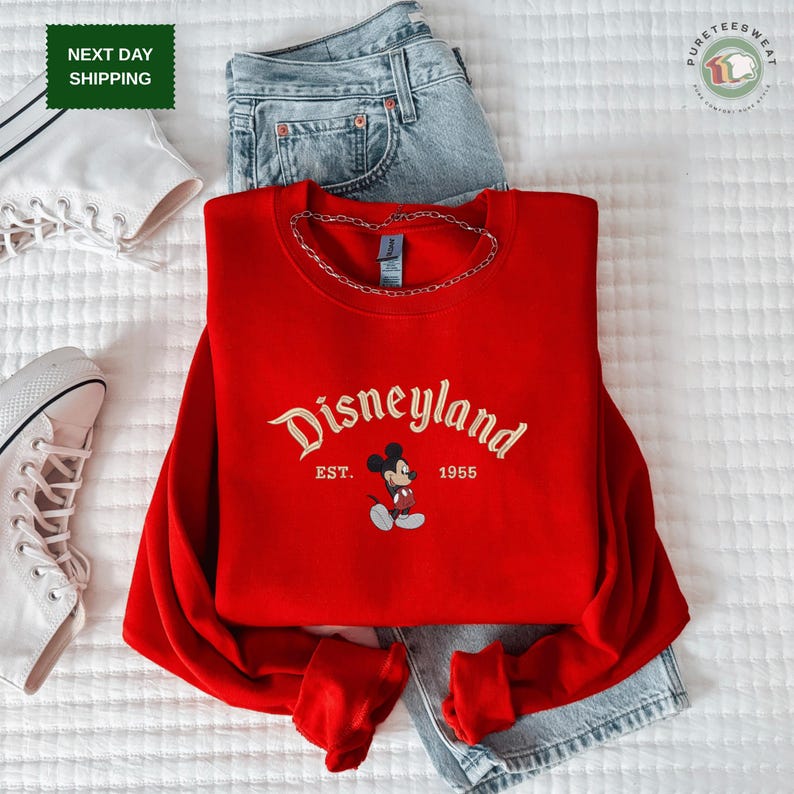 A red Disneyland sweatshirt with the text "Disneyland EST. 1955" printed on it, along with a pair of jeans and white sneakers, all laid out on a white surface.
