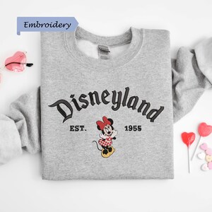 A gray sweatshirt with the text "Disneyland EST. 1955" and a Minnie Mouse graphic, along with a pair of sunglasses and heart-shaped candies, are arranged on a white background.