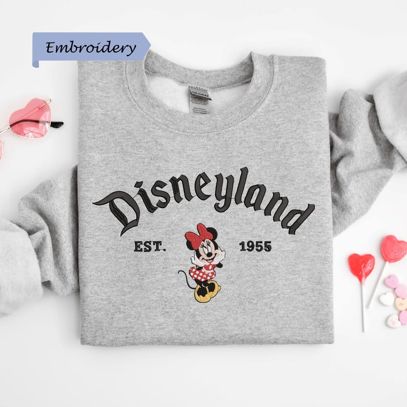 A gray sweatshirt with the text "Disneyland EST. 1955" and a Minnie Mouse graphic, along with a pair of sunglasses and heart-shaped candies, are arranged on a white background.
