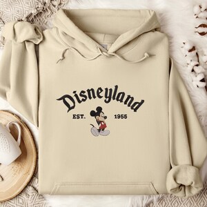 A beige hoodie with the text "Disneyland EST. 1955" and a Mickey Mouse graphic printed on it, along with a wooden tray and some cotton balls in the background.