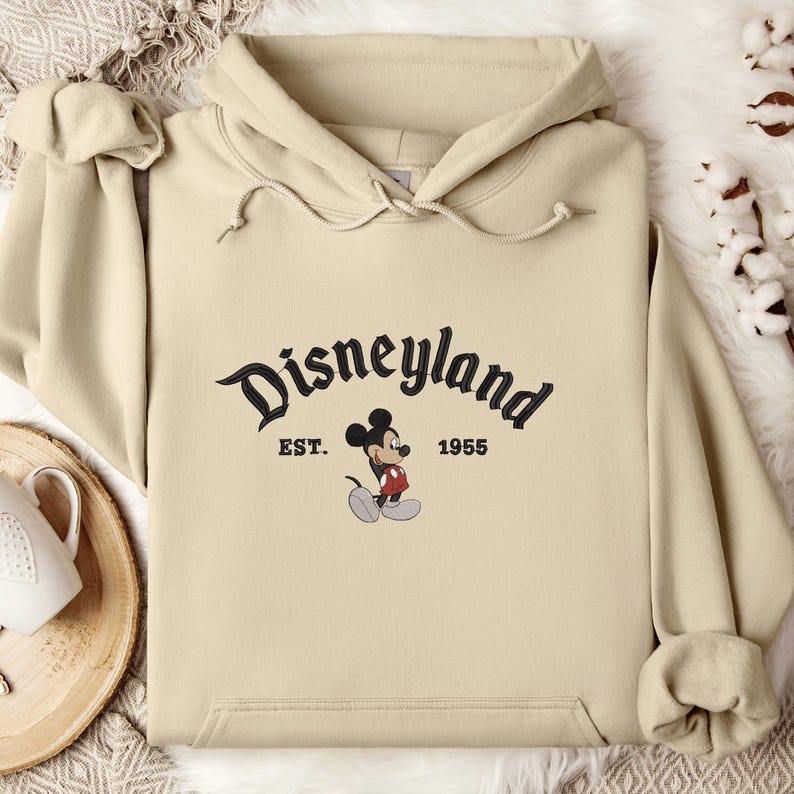 A beige hoodie with the text "Disneyland EST. 1955" and a Mickey Mouse graphic printed on it, along with a wooden tray and some cotton balls in the background.