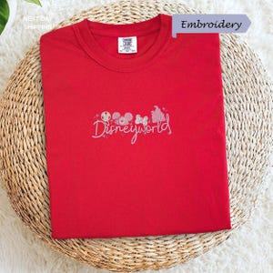 A red t-shirt with the text "Disneyworld" embroidered on it, placed on a woven basket.