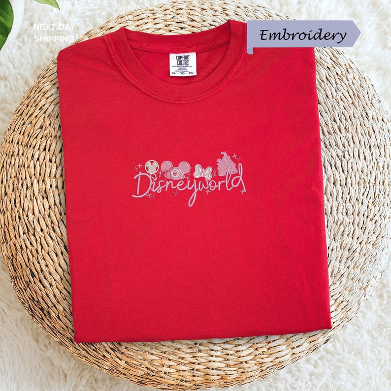 A red t-shirt with the text "Disneyworld" embroidered on it, placed on a woven basket.