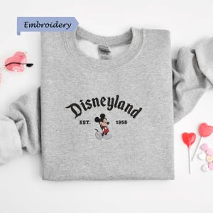 A gray sweatshirt with the Disneyland logo and text, along with some heart-shaped candies and a pair of sunglasses, all laid out on a white background.