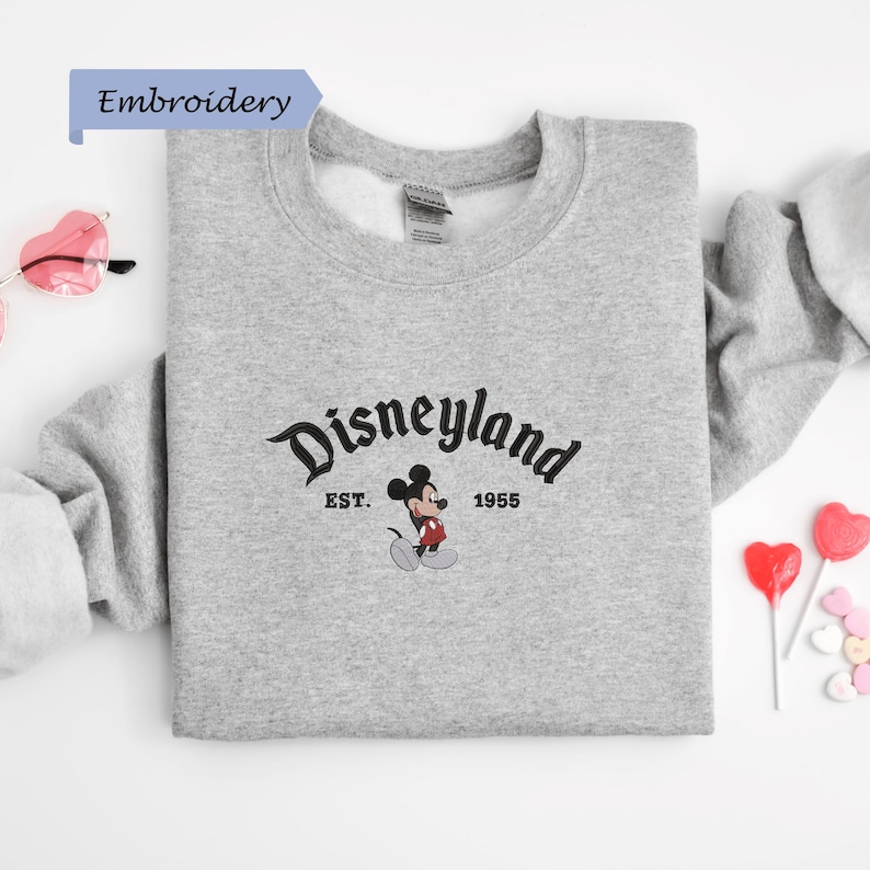 A gray sweatshirt with the Disneyland logo and text, along with some heart-shaped candies and a pair of sunglasses, all laid out on a white background.