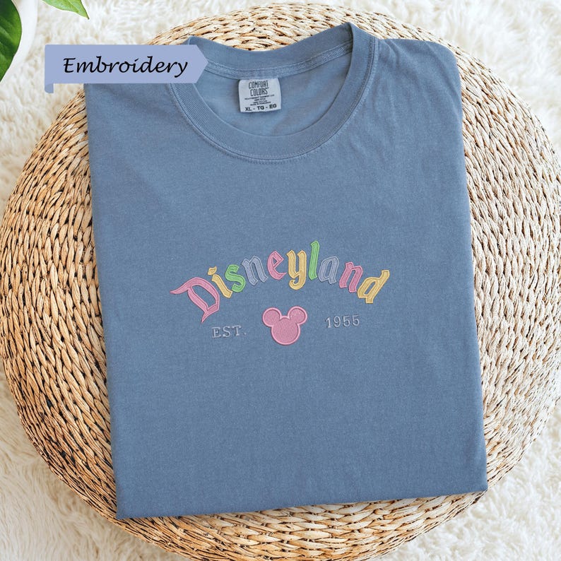 A blue t-shirt with the text "Disneyland EST. 1955" embroidered on it, along with a Mickey Mouse logo, is displayed on a woven basket.
