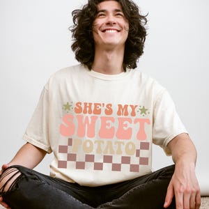 Può includere: T-shirt color avorio con la scritta "SHE'S MY SWEET POTATO" in rosa, pesca e beige. Il testo è sopra un motivo a scacchi marrone e beige. La persona indossa jeans neri.