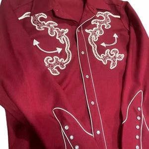 Puede incluir: Una camisa de manga larga de color burdeos con bordados blancos y ribetes blancos. La camisa tiene un cierre de botones en la parte delantera y un cuello puntiagudo.