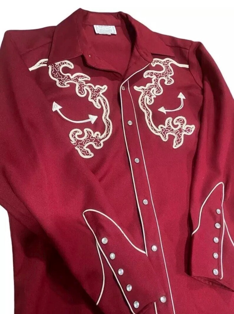 Puede incluir: Una camisa de manga larga de color burdeos con bordados blancos y ribetes blancos. La camisa tiene un cierre de botones en la parte delantera y un cuello puntiagudo.
