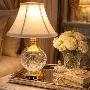 May include: A bedside table arrangement featuring a crystal and gold table lamp with a cream lampshade. A mirrored tray holds a vase of white roses, a perfume bottle, and a book with a pearl necklace. The lamp has a round, cut-glass base.