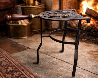 May include: A black metal trivet with a wooden handle, set in front of a fireplace. The trivet has a decorative, openwork design and is supported by three legs. A lit candle and brass containers are in the background.
