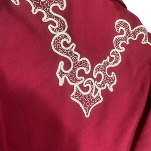Puede incluir: Una camisa color burdeos con un dise&ntilde;o bordado blanco en la espalda. El dise&ntilde;o es un patr&oacute;n floral estilizado.