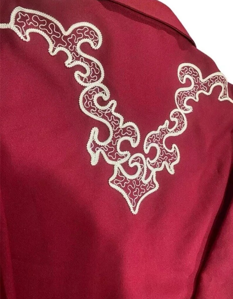 Puede incluir: Una camisa color burdeos con un dise&ntilde;o bordado blanco en la espalda. El dise&ntilde;o es un patr&oacute;n floral estilizado.