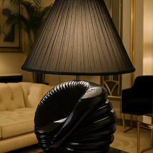 May include: A black table lamp with a pleated lampshade and a sculptural base. The base has a glossy finish and a spiral design. The lamp is on a dark surface, with a sofa and artwork in the background.